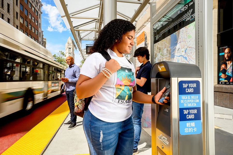 A youth holds their Android phone up to a standing Clipper card reader located at an outdoor bus stop, using Clipper on their phone to pay for transit.