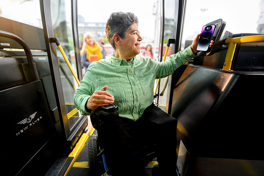 An adult in a wheelchair boards a bus, holding an iPhone up to Clipper card reader, using Clipper on their phone to pay for transit; a “Done” screen is displayed on the phone.