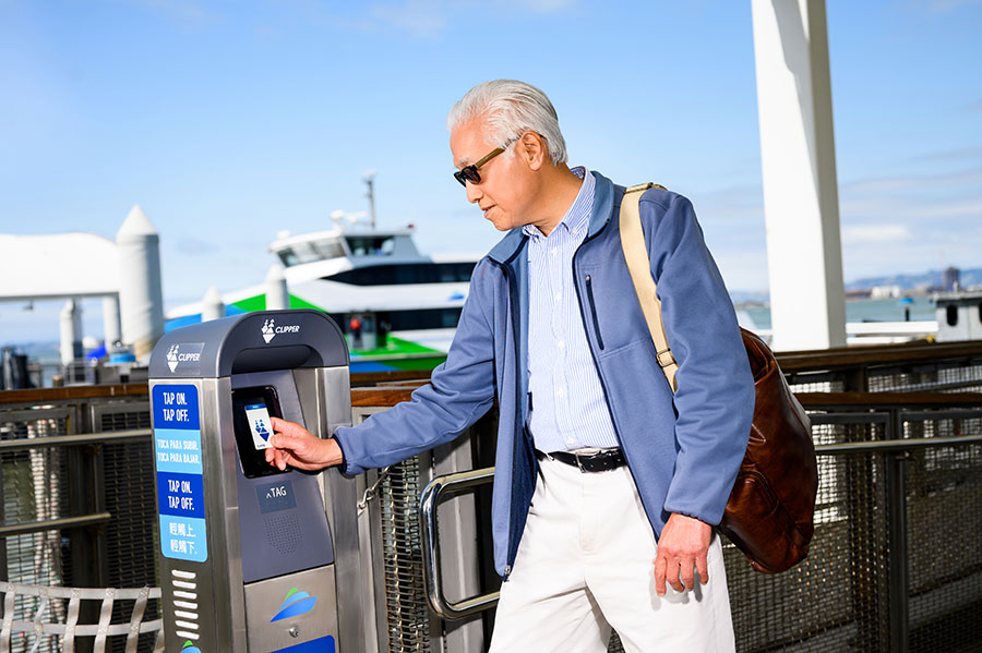 A senior walking up to a standing Clipper card reader at the San Francisco Bay Ferry, holding a Senior Clipper card up to the reader to pay for transit.