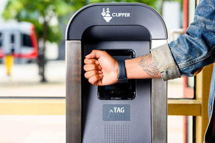 A close-up of a person's hand in front of a standing Clipper card reader, holding their Apple Watch up to the reader to pay for transit with a mobile Clipper card.