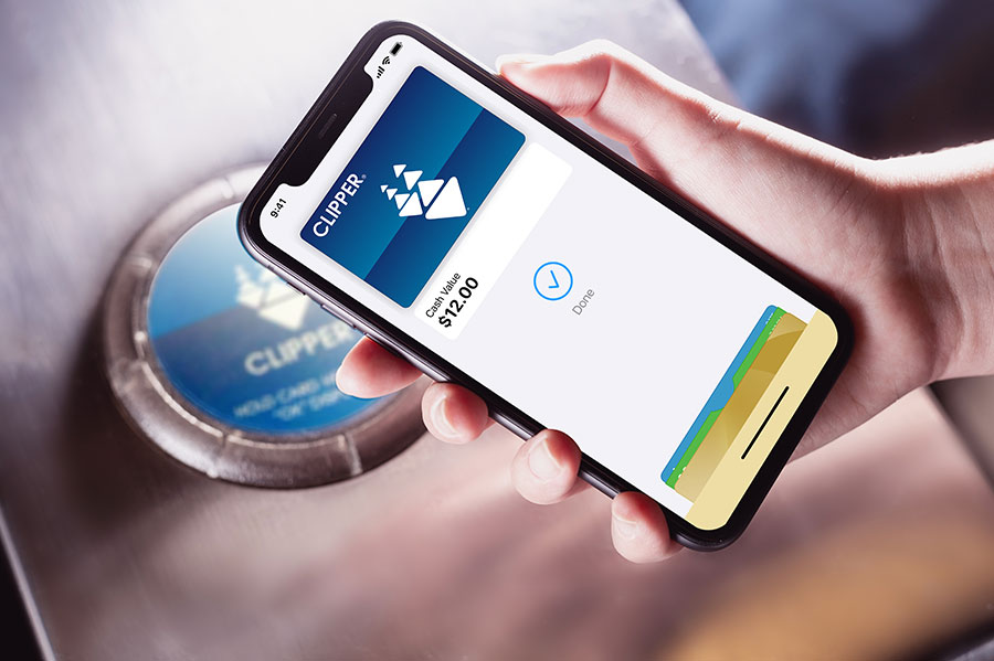 Close-up of a hand holding an iPhone over a BART card reader target, using Clipper on their phone to pay for transit; a “Done” screen is displayed on the phone.