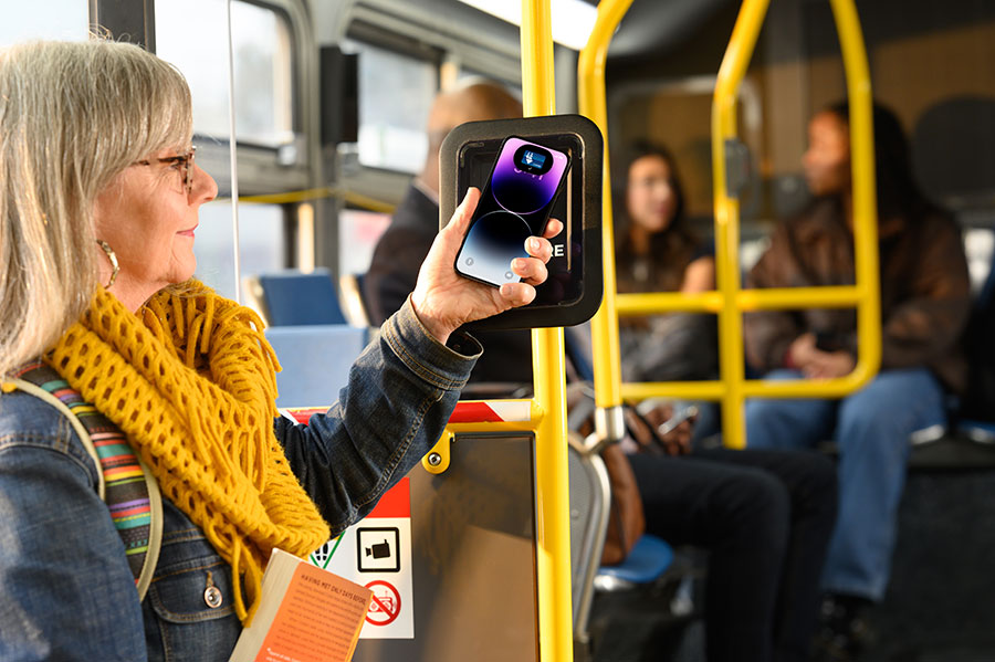 An adult boards a bus, holding their iPhone up to Clipper bus card reader, using Clipper on her phone to pay for transit; a “Done” screen is displayed on the phone.